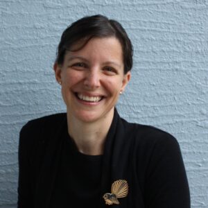 Image of Rebecca Travaglia - a Pākehā woman with short dark hair, smiling at the camera, wearing a black top and a piwakaka (fantail) brooch.