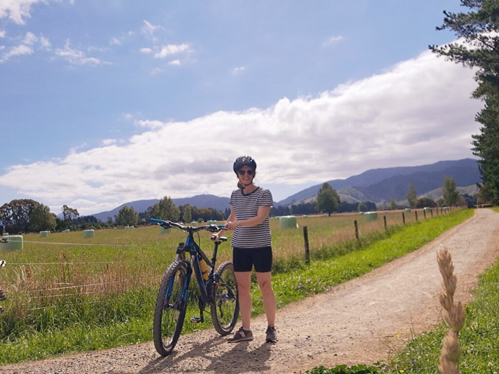 An image of Pip in bike helmet standing next to her bike on the Greytown Rail Trail on a sunny day, with the Tararuas in the distance
